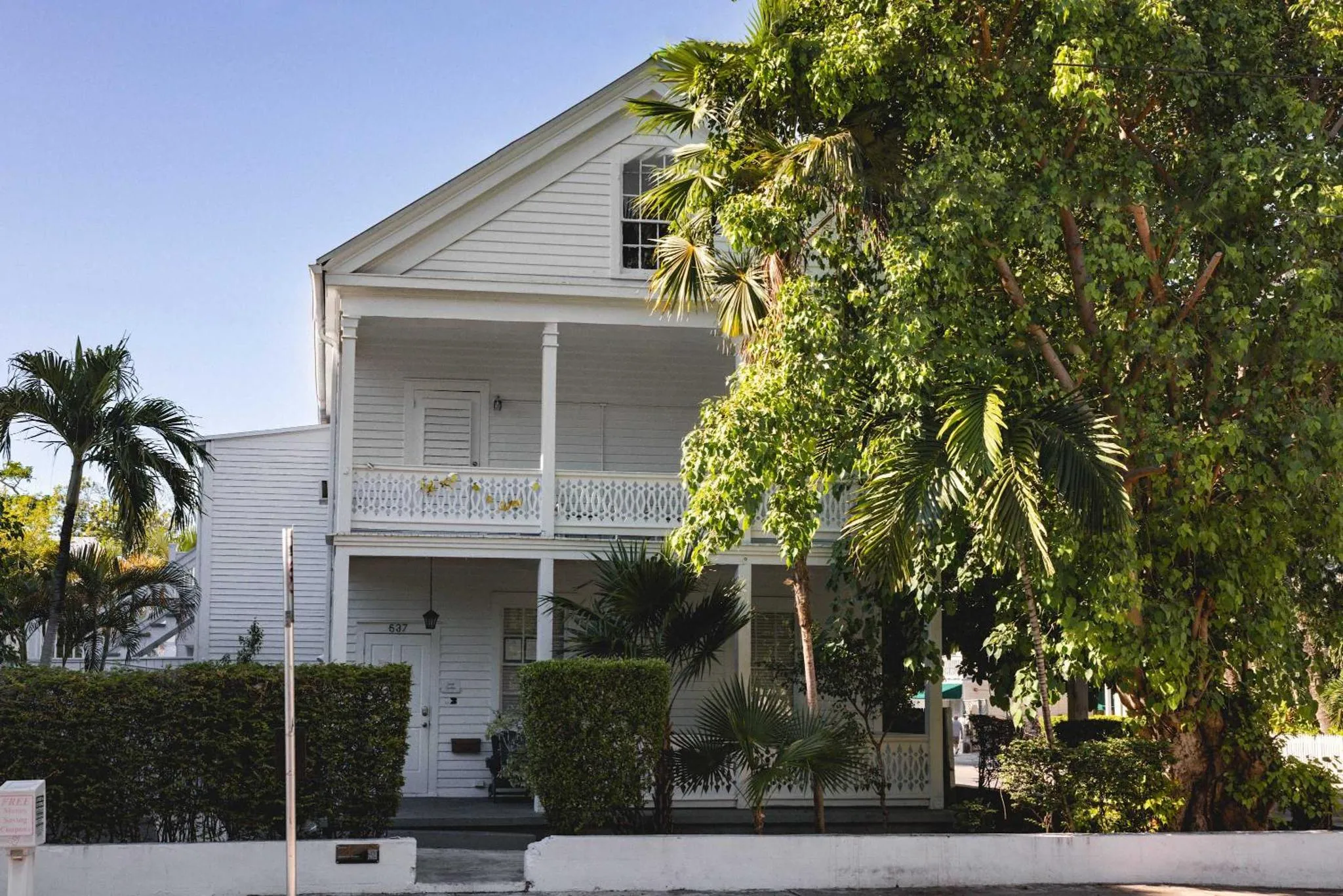 Maison blanche à deux étages avec une véranda ombragée par des palmiers.