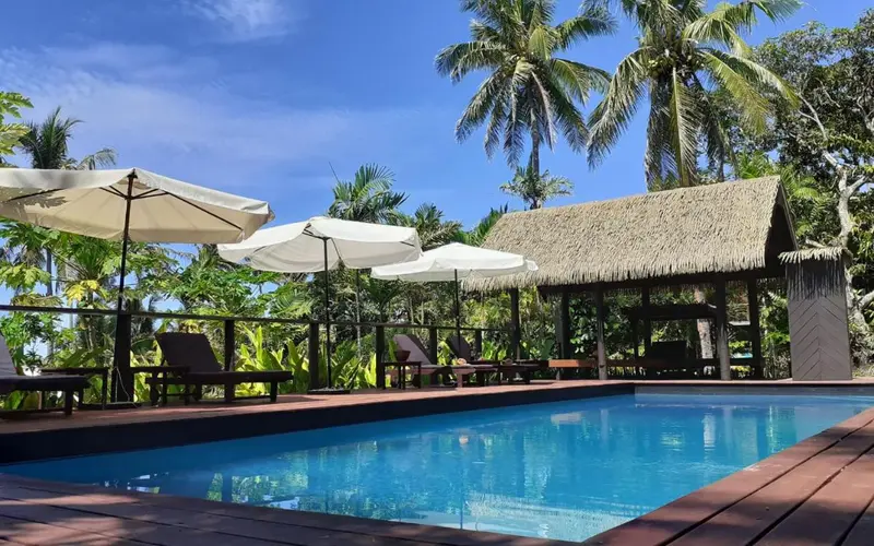 Une piscine bordée d'une terrasse avec des transats et des parasols, entourée de végétation tropicale.