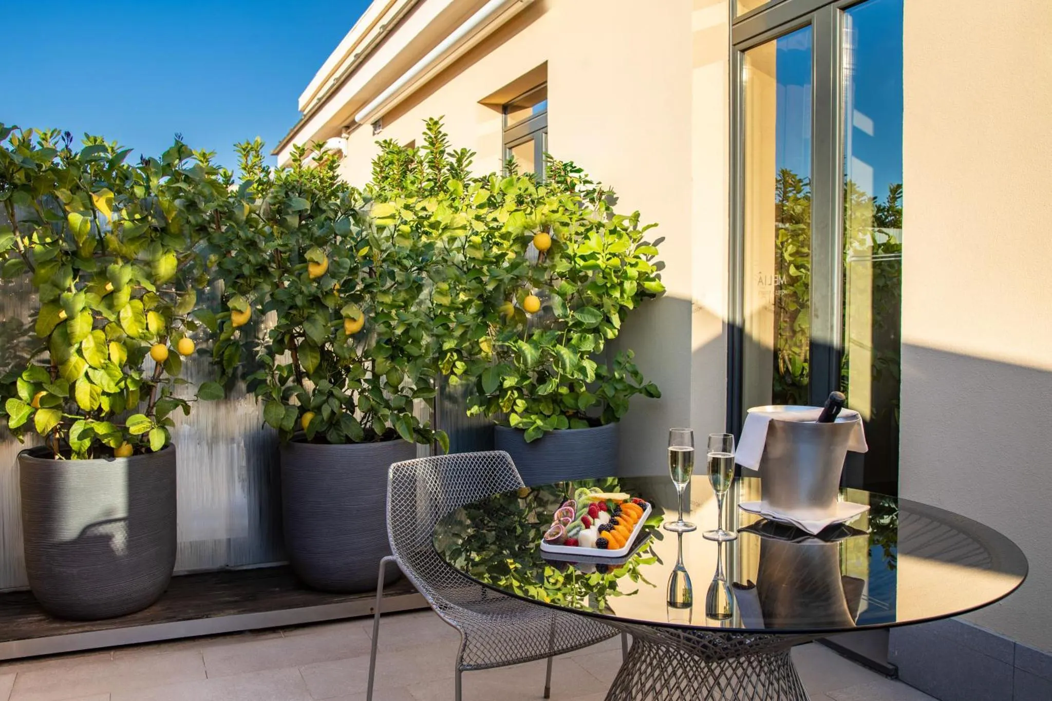 Une table ronde en verre avec des fruits et une bouteille fraîche sur une terrasse ensoleillée.