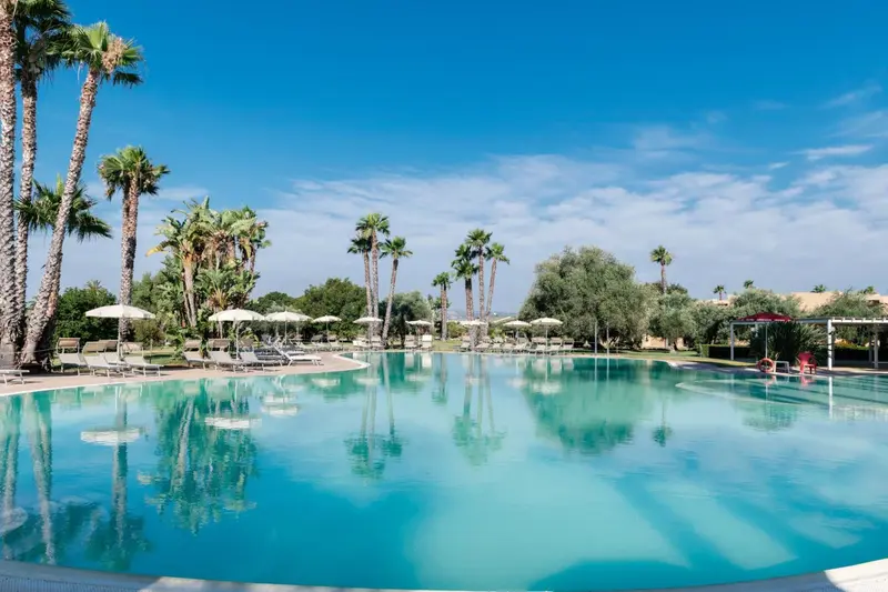 Une piscine turquoise reflète les palmiers sous un ciel bleu nuageux.