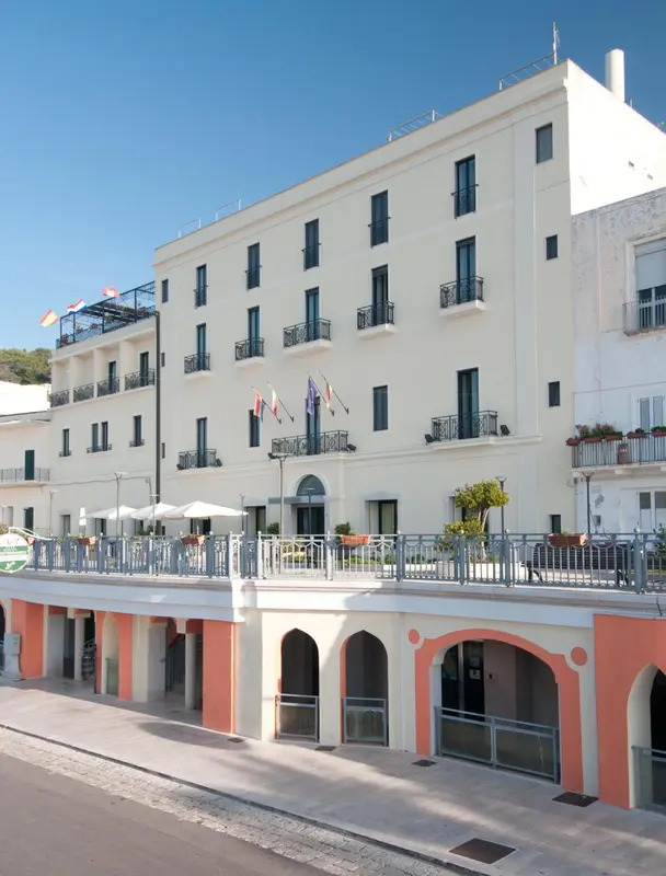 Bâtiment blanc à plusieurs étages avec des balcons et une entrée ombragée.