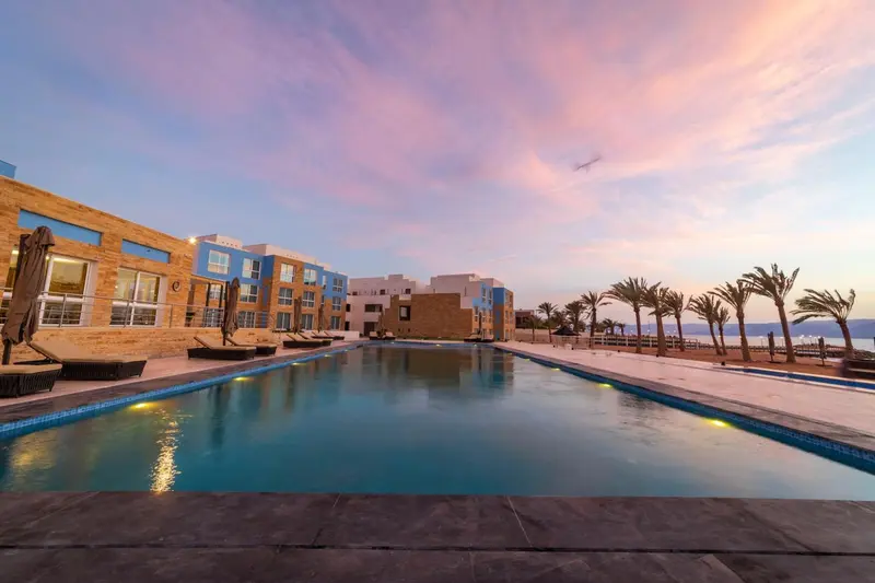 Une piscine rectangulaire bordée de transats devant un bâtiment moderne au coucher du soleil.