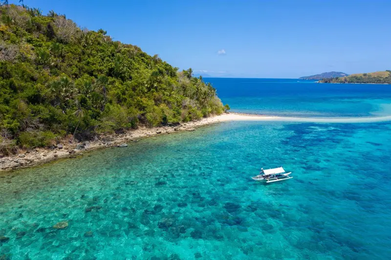 Une petite embarcation navigue sur une eau turquoise peu profonde près d'une plage de sable blanc.