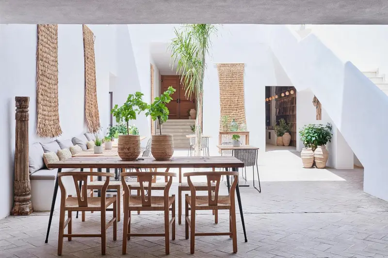 Une table en bois est entourée de chaises dans un espace extérieur lumineux et décoré de plantes.