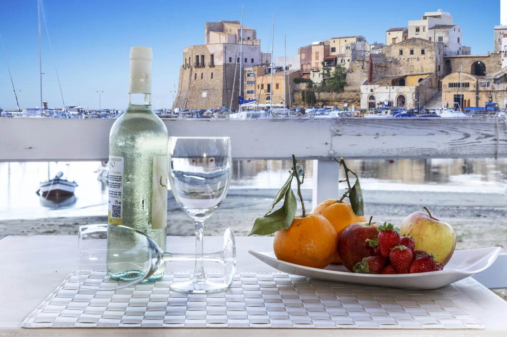 Nature morte avec une bouteille de vin, un verre, des fruits et une vue sur un port.