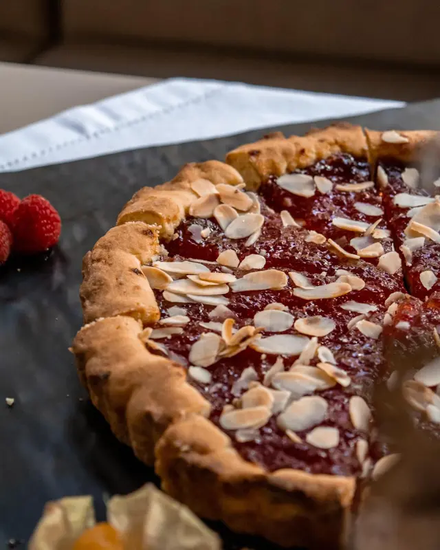 Une tarte aux amandes et aux framboises est présentée sur une surface sombre.