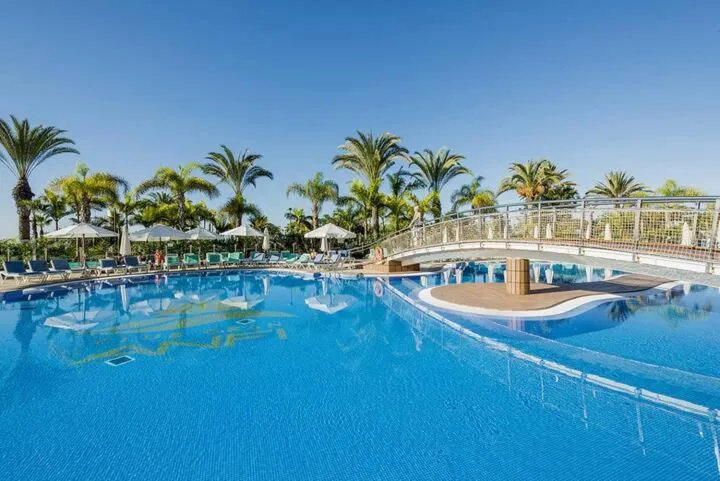 Une piscine bleue bordée de transats et de palmiers sous un ciel clair.