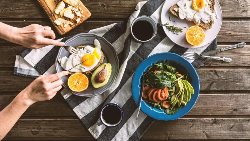 Une table en bois présente des assiettes garnies d'œufs, d'avocat et de salade, accompagnées de tasses de café.