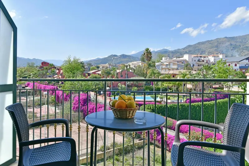 Une table ronde avec des fruits est entourée de chaises sur une terrasse fleurie avec vue sur un paysage montagneux.