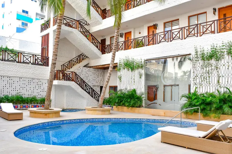 Une piscine circulaire est entourée de chaises longues et de végétation tropicale devant un bâtiment blanc.