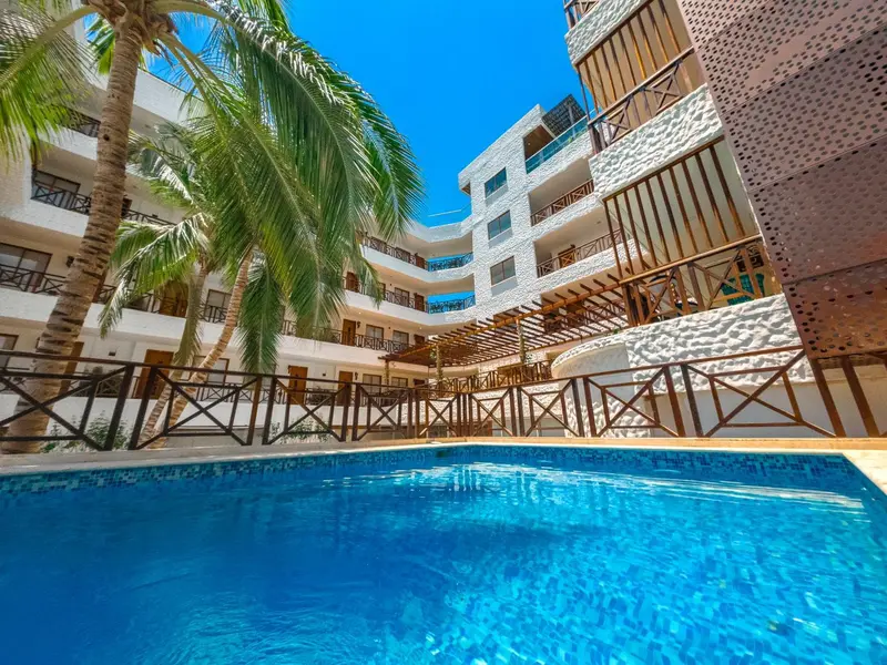 Vue d'une piscine turquoise devant un bâtiment blanc et beige avec des balcons.