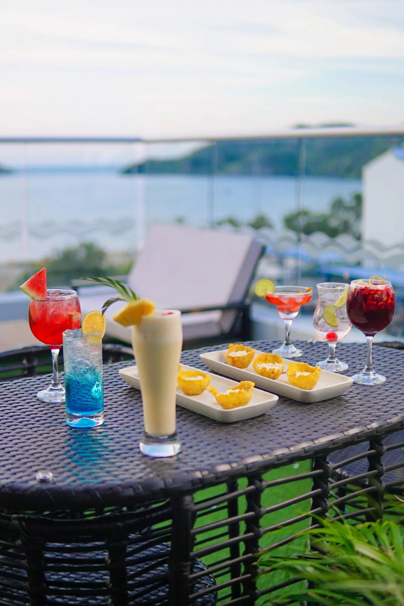 Une table basse en osier présente des cocktails colorés et des amuse-gueules sur une terrasse avec vue sur la mer.