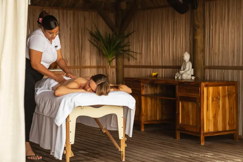 Une femme reçoit un massage sur une table en bois dans un espace extérieur ombragé.