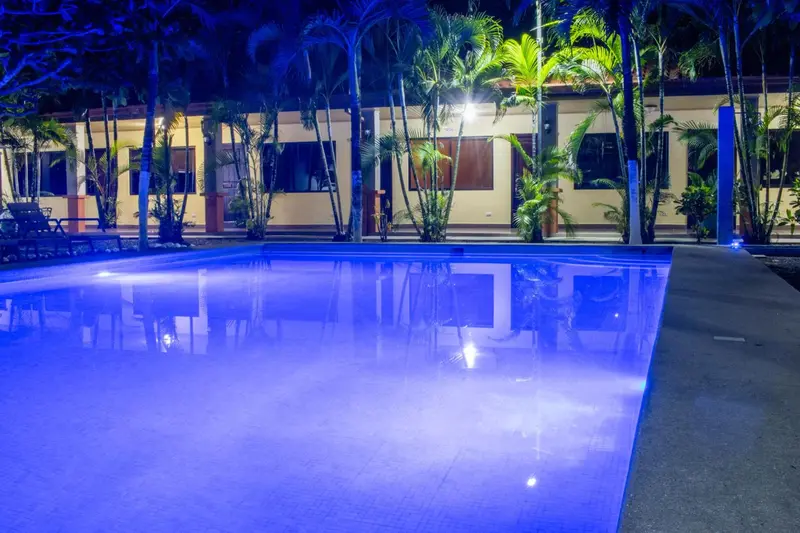 Piscine illuminée en bleu sous un ciel nocturne, avec des bâtiments en arrière-plan.