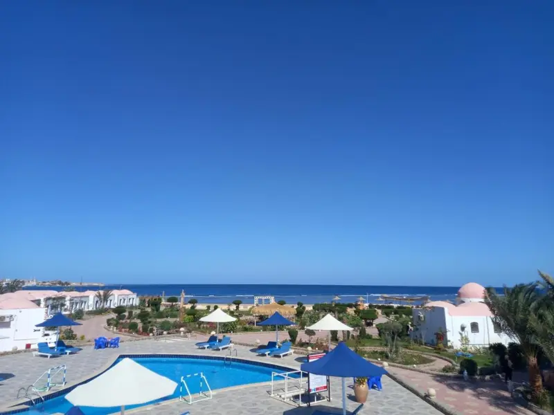Piscine turquoise bordée de transats sous des parasols blancs et bleus, avec vue sur la mer.