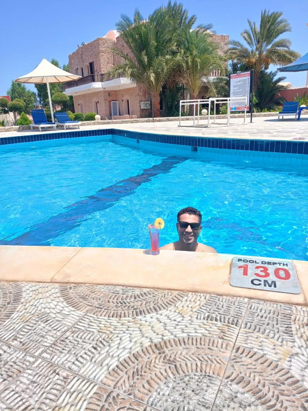 Une piscine bleue avec un homme souriant et un cocktail sur le bord.
