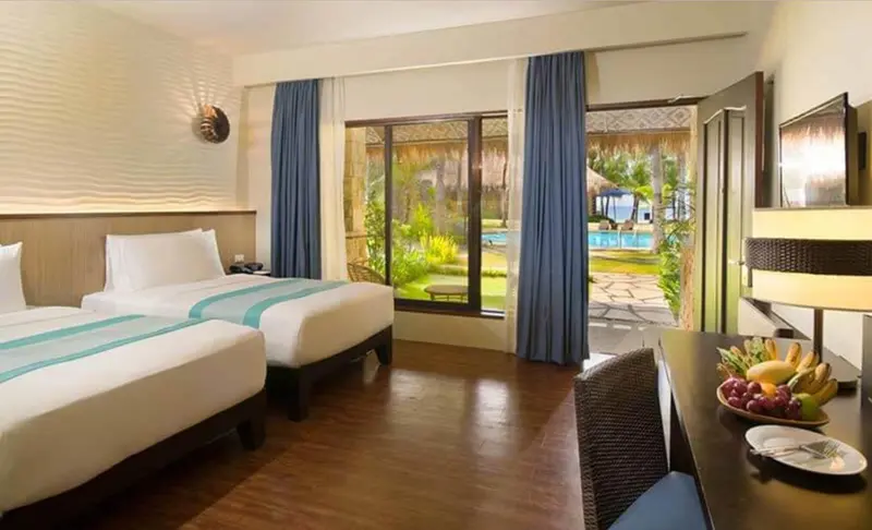 Chambre lumineuse avec deux lits et vue sur une piscine ensoleillée.