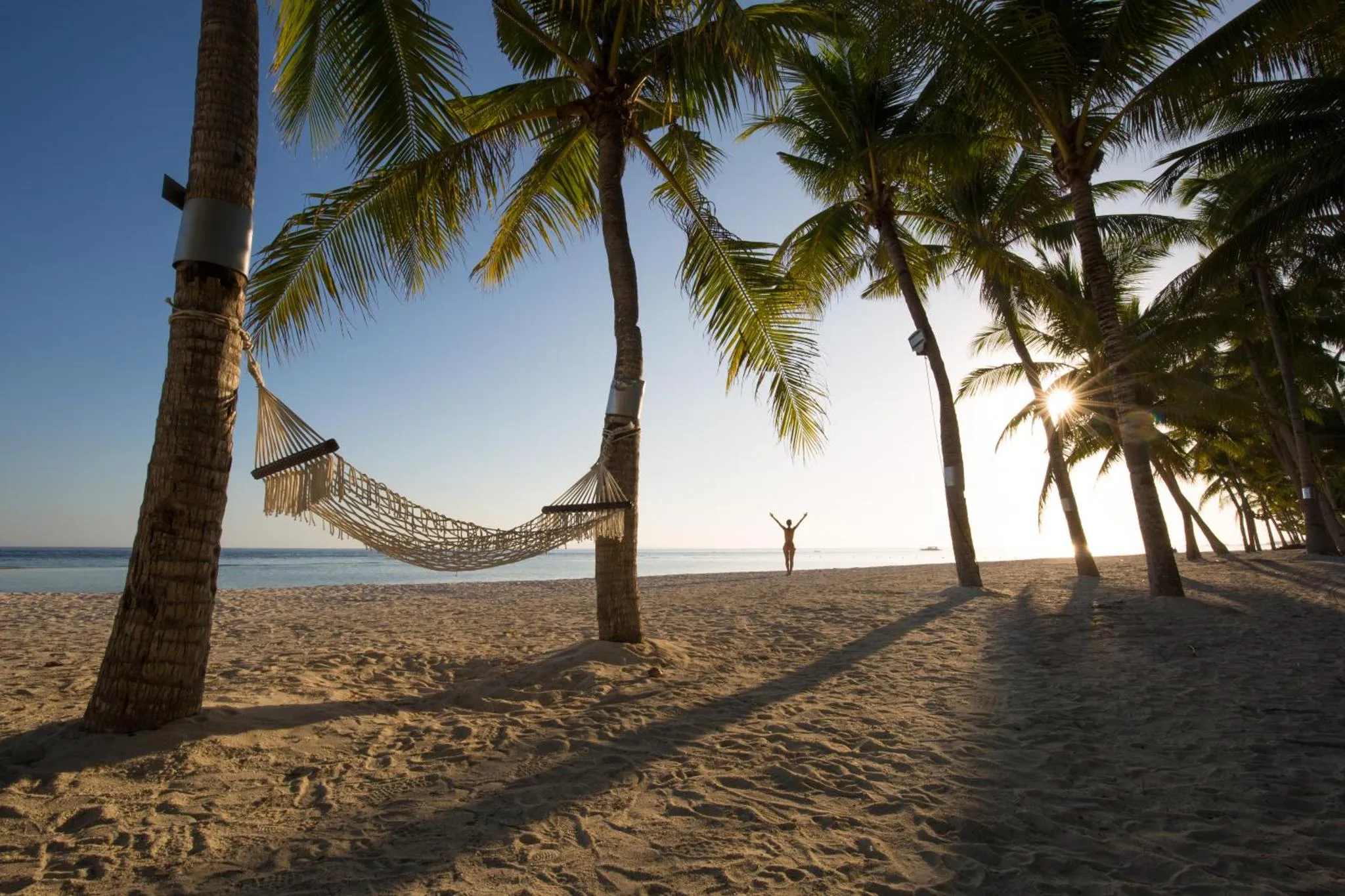 Une personne se tient sur une plage de sable fin au coucher du soleil, avec des palmiers et un hamac.