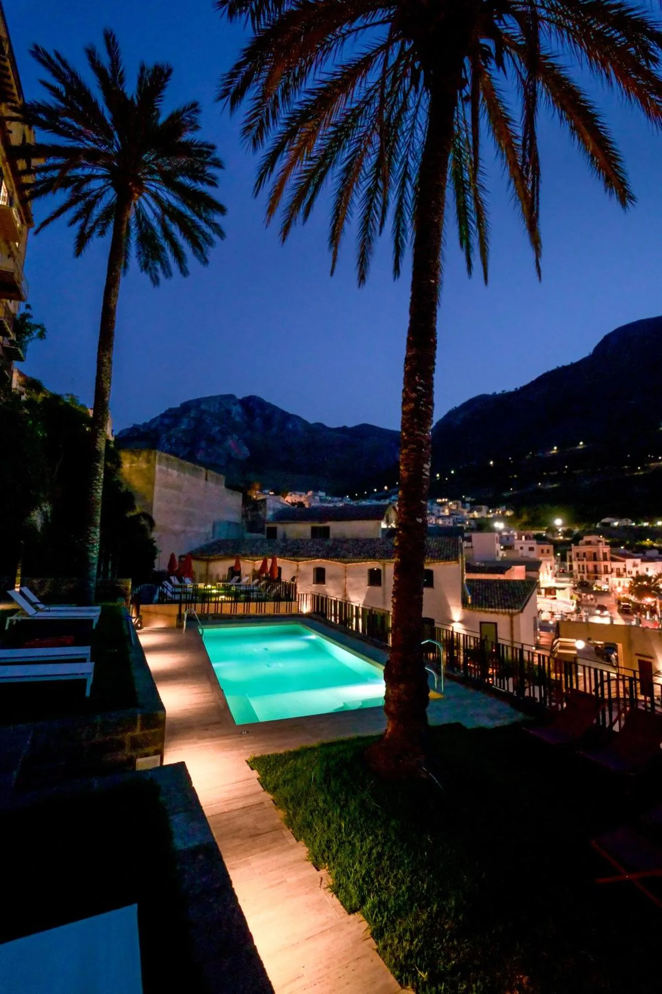 Une piscine illuminée au crépuscule, avec des palmiers et des montagnes en arrière-plan.