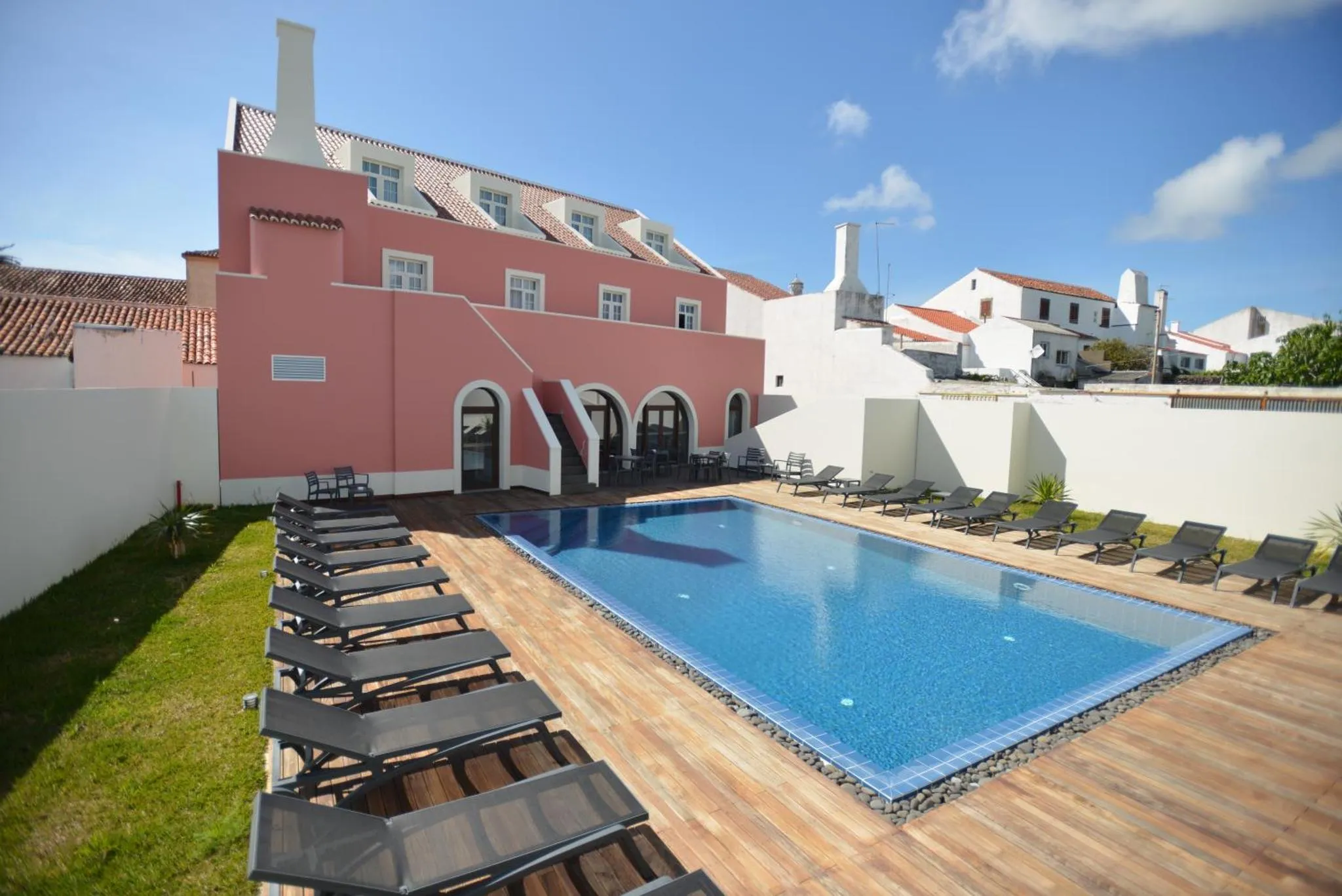 Piscine rectangulaire entourée de transats dans un jardin soigné devant un bâtiment rose.