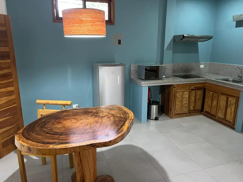 Une table en bois massif se trouve dans une pièce aux murs bleus, équipée d'une kitchenette.
