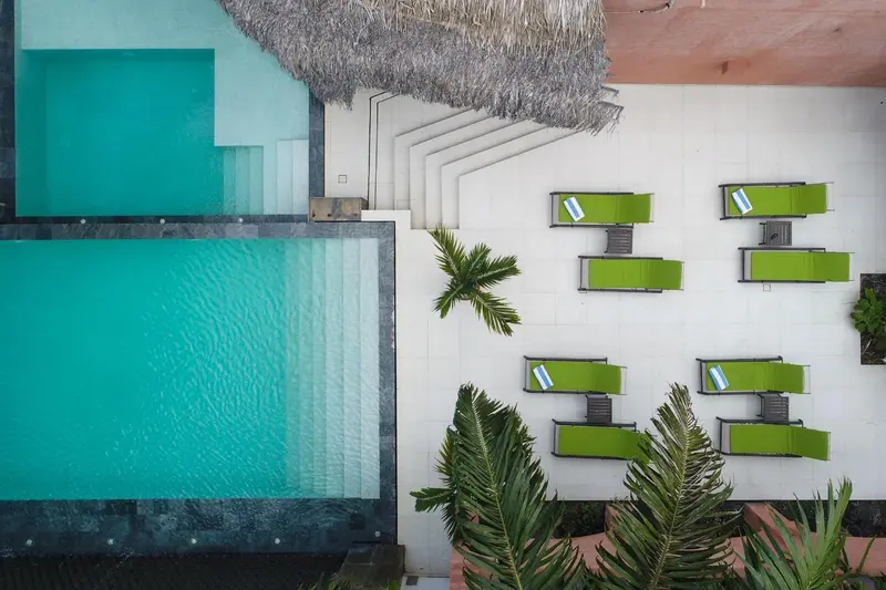Vue aérienne d'une piscine rectangulaire entourée de chaises longues vertes et de végétation tropicale.