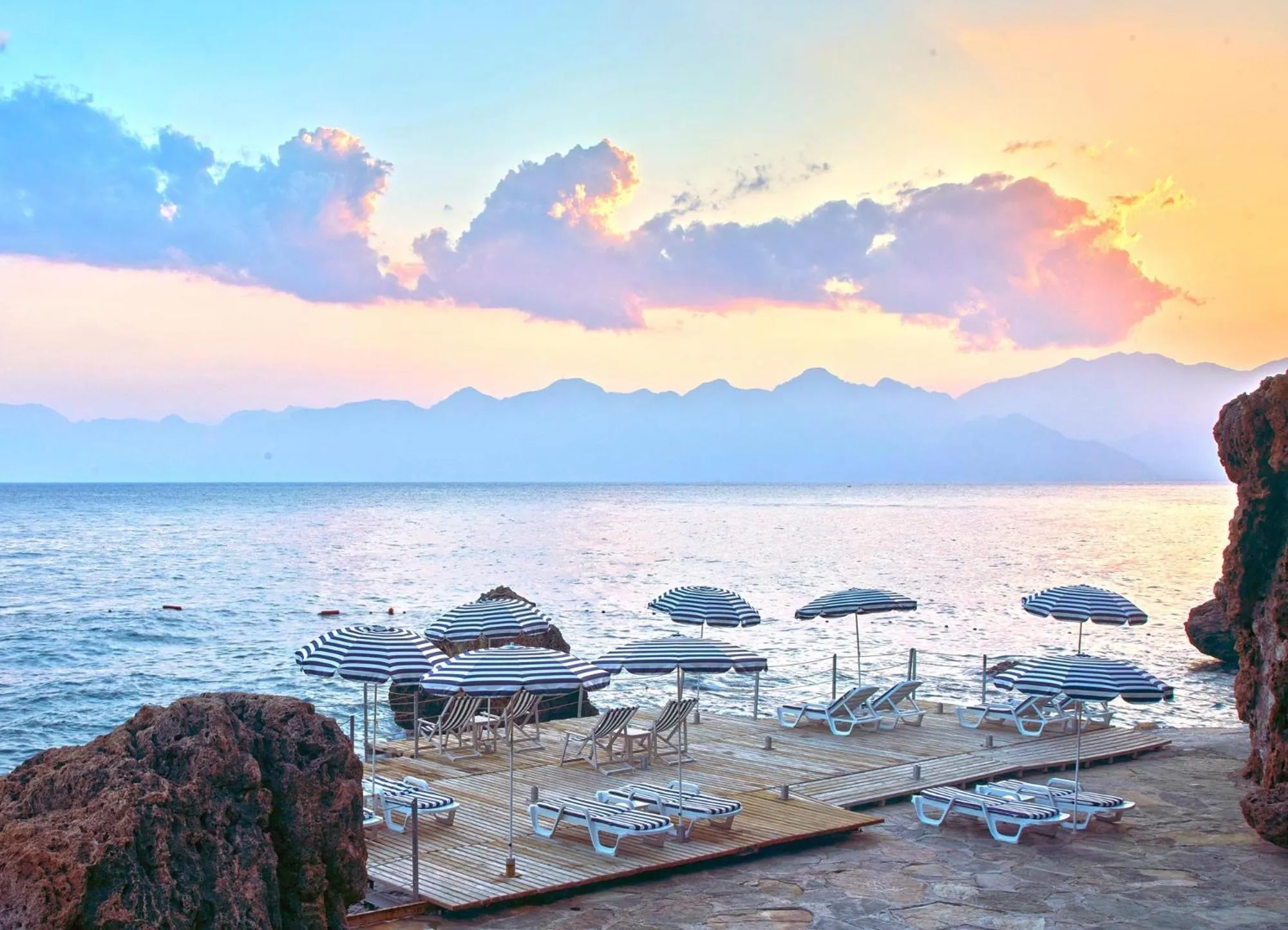 Transats et parasols sur une plage au coucher du soleil avec vue sur la mer et les montagnes.