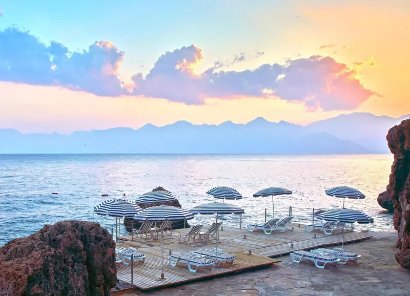 Transats et parasols sur une plage au coucher du soleil avec vue sur la mer et les montagnes.