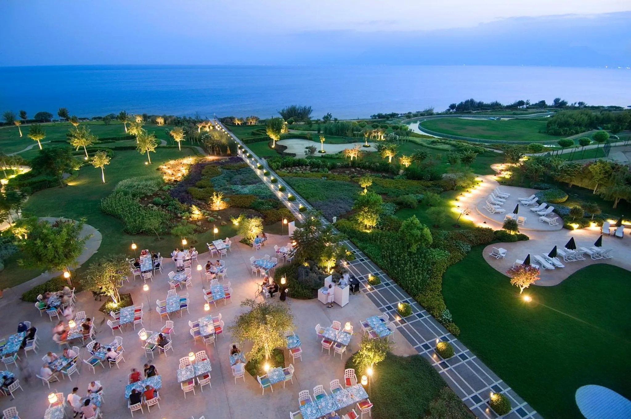 Vue aérienne d'un restaurant en plein air illuminé au crépuscule, avec des tables et des chaises disposées sur une pelouse verdoyante.