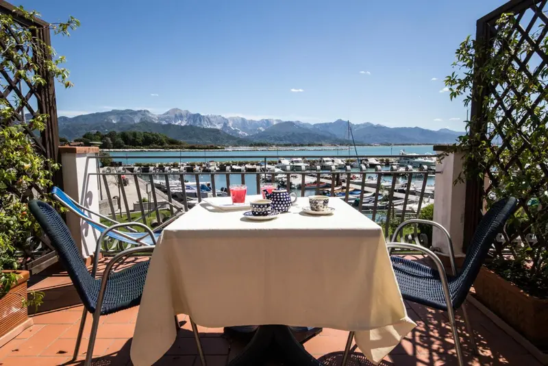 Une table est dressée pour un repas sur une terrasse ensoleillée avec vue sur la mer et les montagnes.