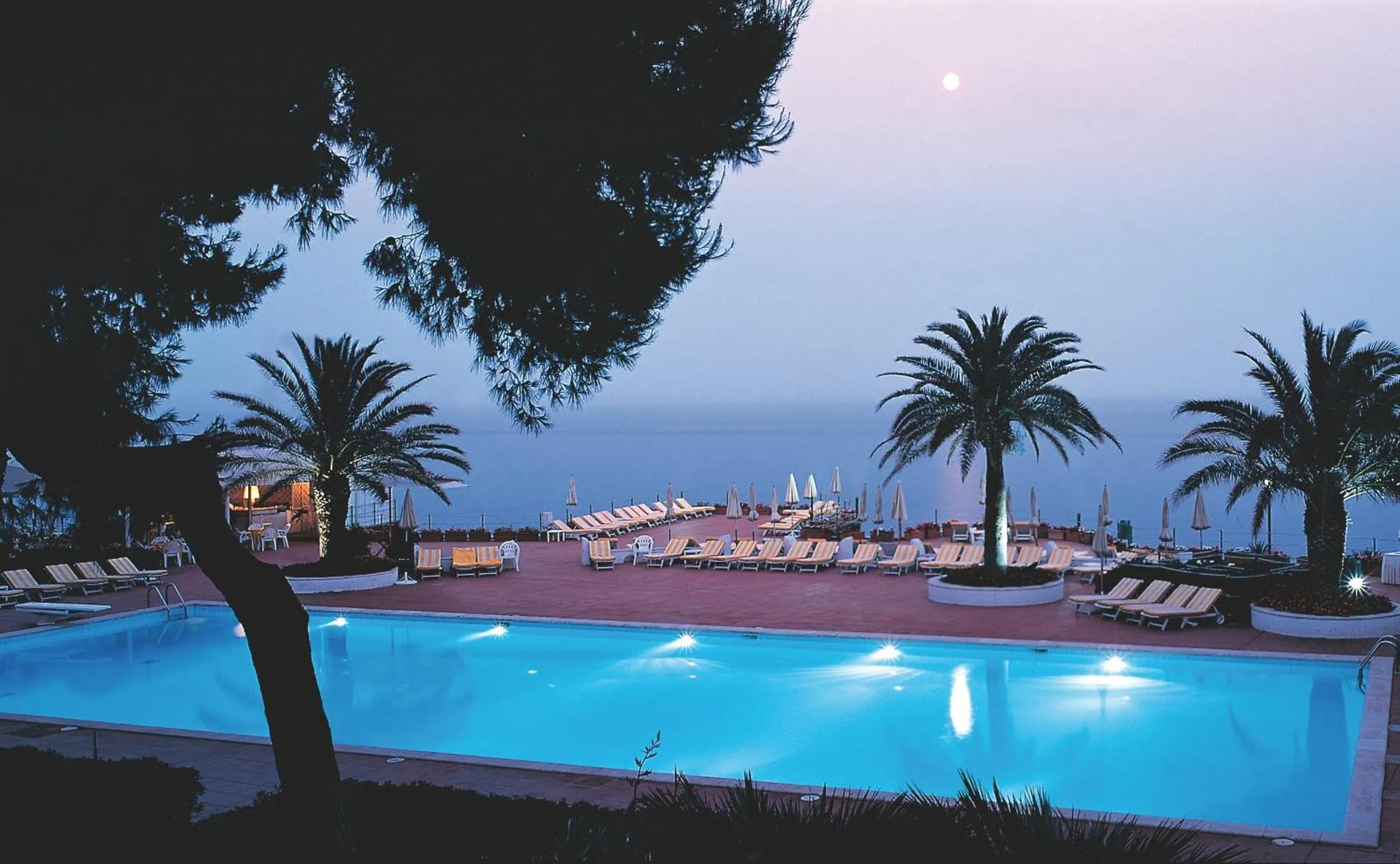 Une piscine illuminée au premier plan, avec des transats et des palmiers en arrière-plan.