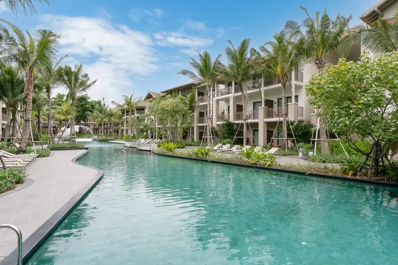 Une piscine turquoise sinueuse bordée de chaises longues, devant un bâtiment beige à plusieurs étages.