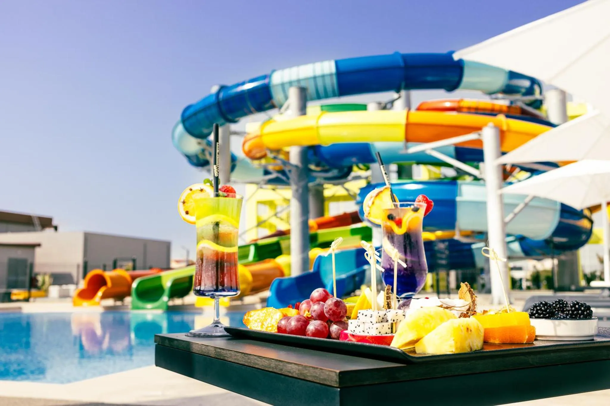 Des cocktails colorés et des fruits frais sont présentés sur une table basse au bord d'une piscine avec toboggans.
