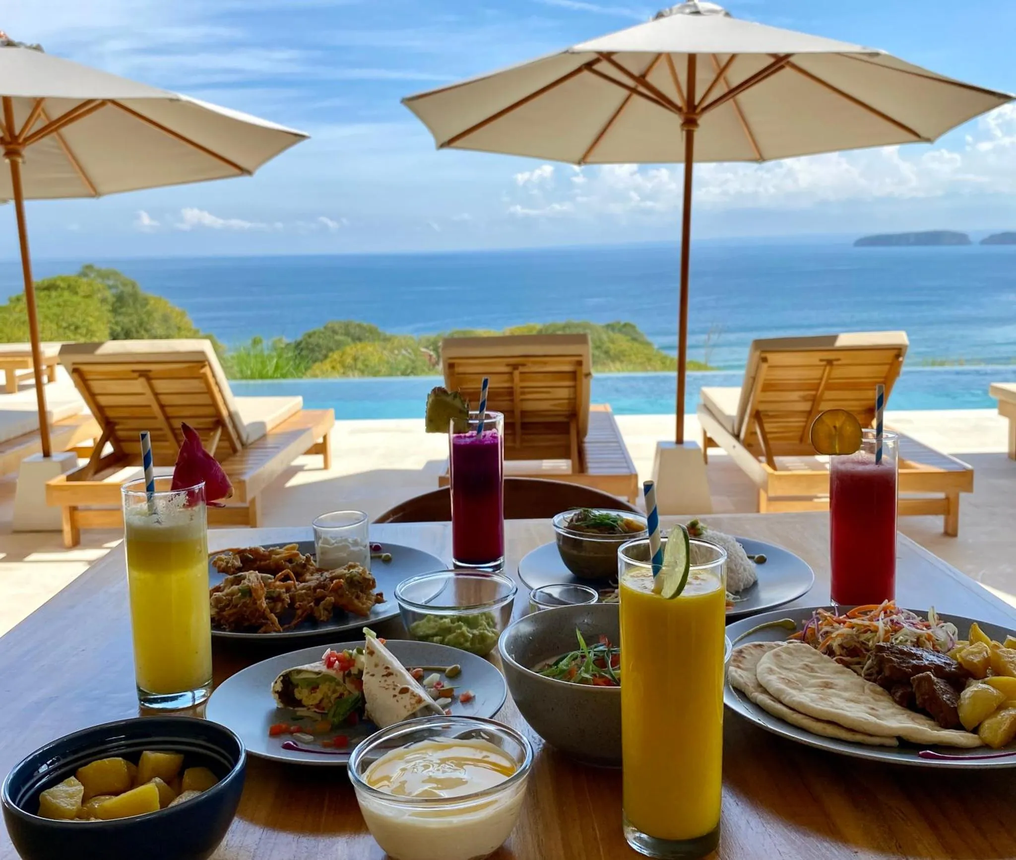 Une table est dressée avec des plats colorés et des boissons fraîches, face à une piscine à débordement et une vue sur la mer.