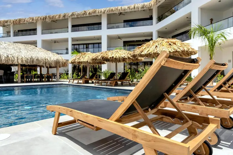 Transats en bois au bord d'une piscine ensoleillée, ombragés par des parasols en chaume.
