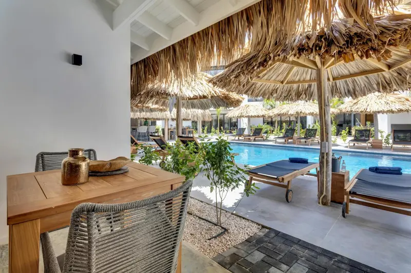 Une table en bois est mise en scène devant une piscine bordée de chaises longues sous des toits de chaume.