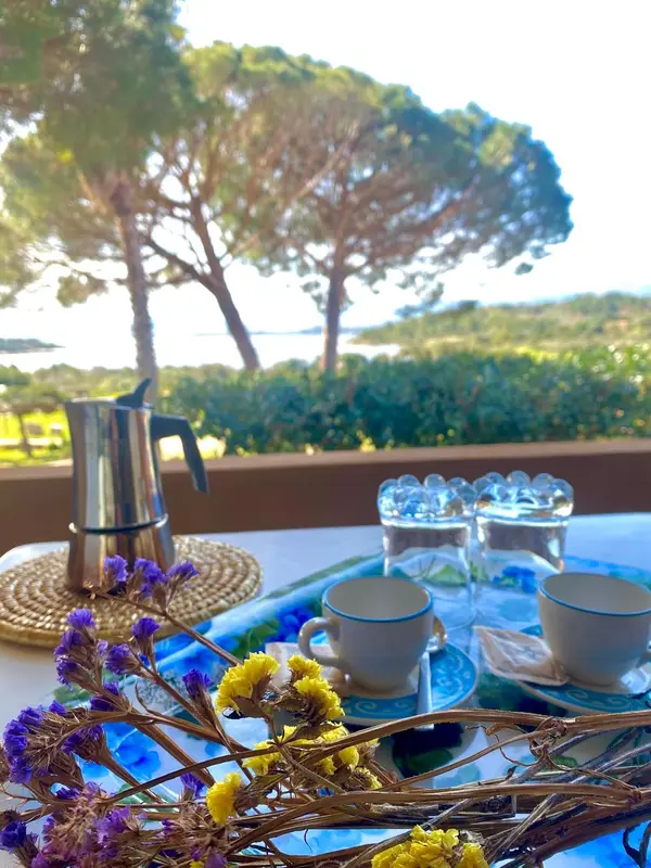 Une table est dressée avec une cafetière, des tasses et des fleurs sur une terrasse.