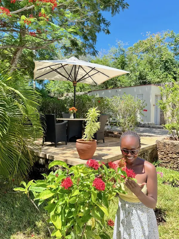 Une femme souriante se tient près d'arbustes en fleurs dans un jardin ensoleillé.