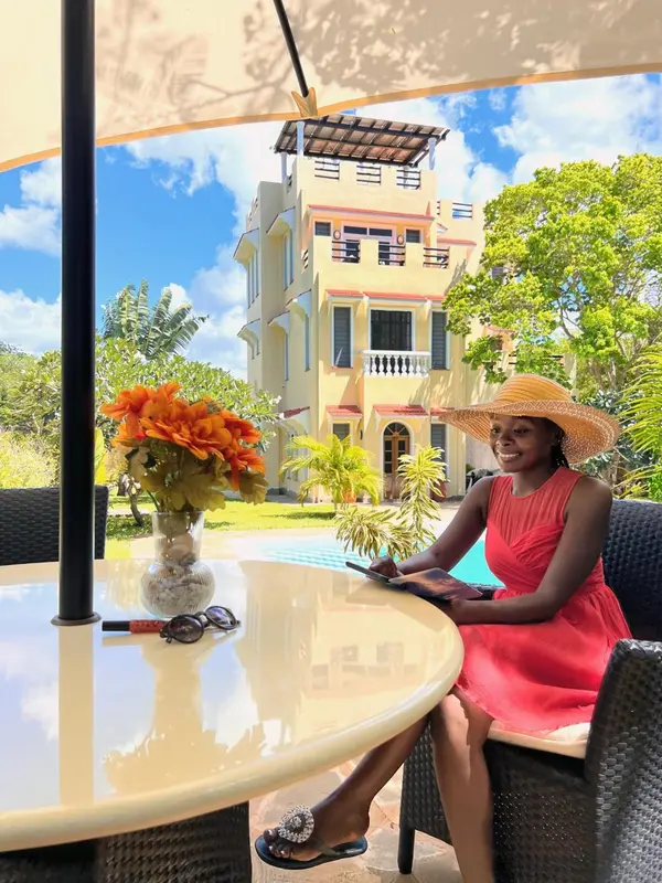 Une femme souriante est assise à une table en extérieur devant un bâtiment blanc.