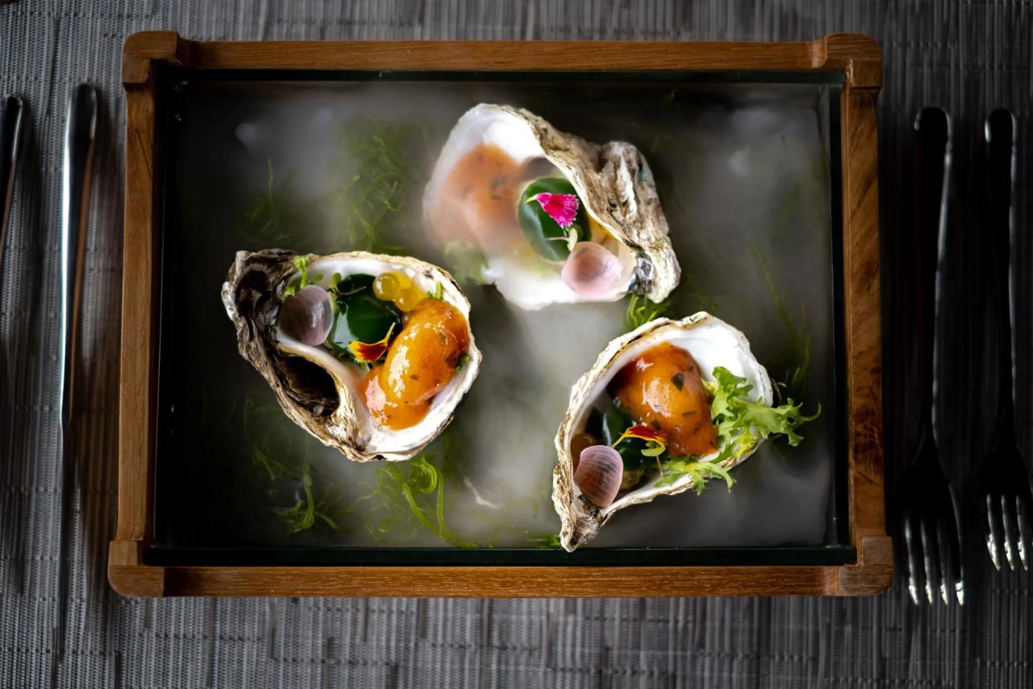 Trois huîtres fraîches, décorées de caviar et de fleurs, sont présentées sur un plateau noir.