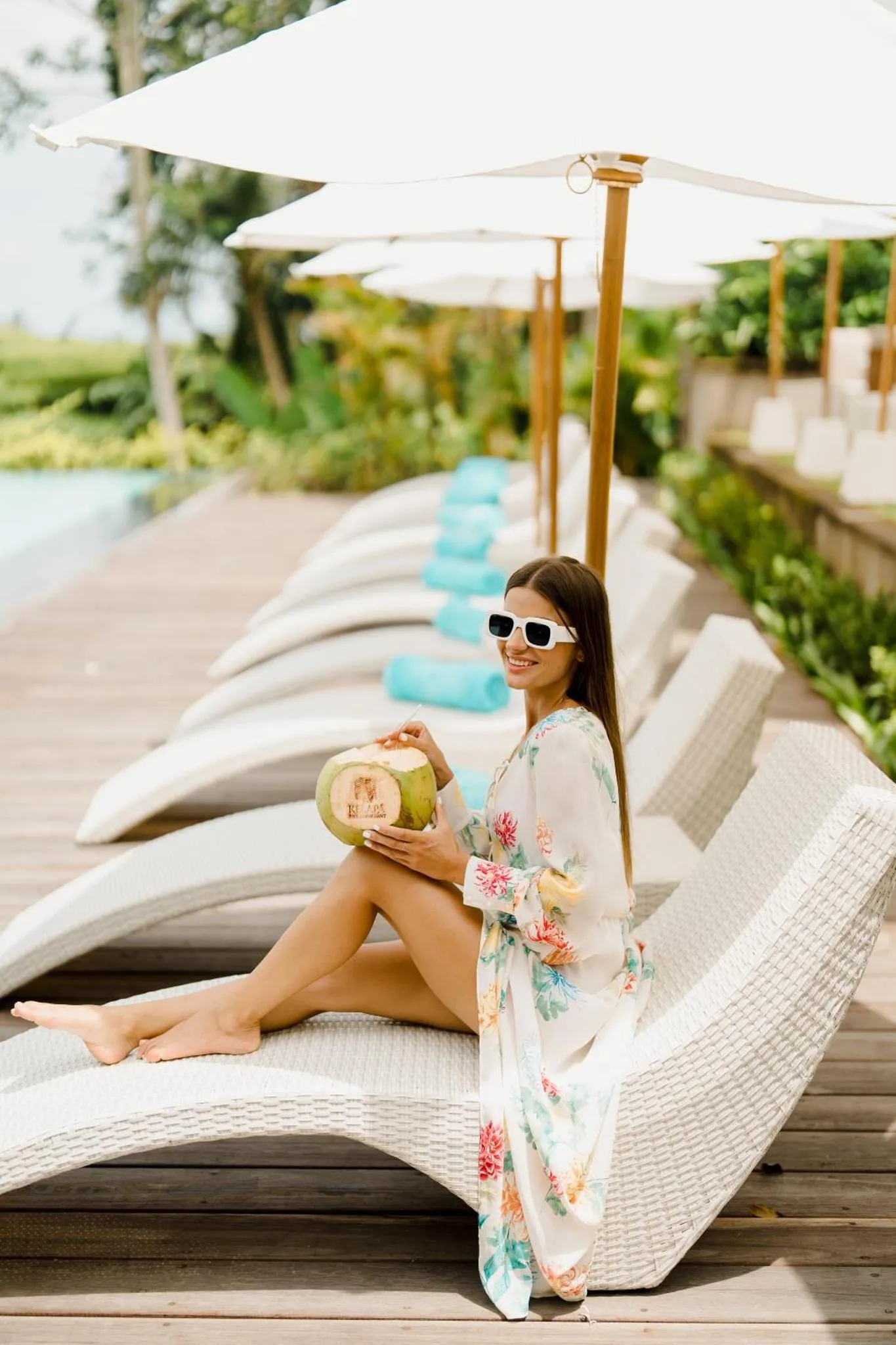 Une femme assise sur un transat au bord d'une piscine, tenant une noix de coco.