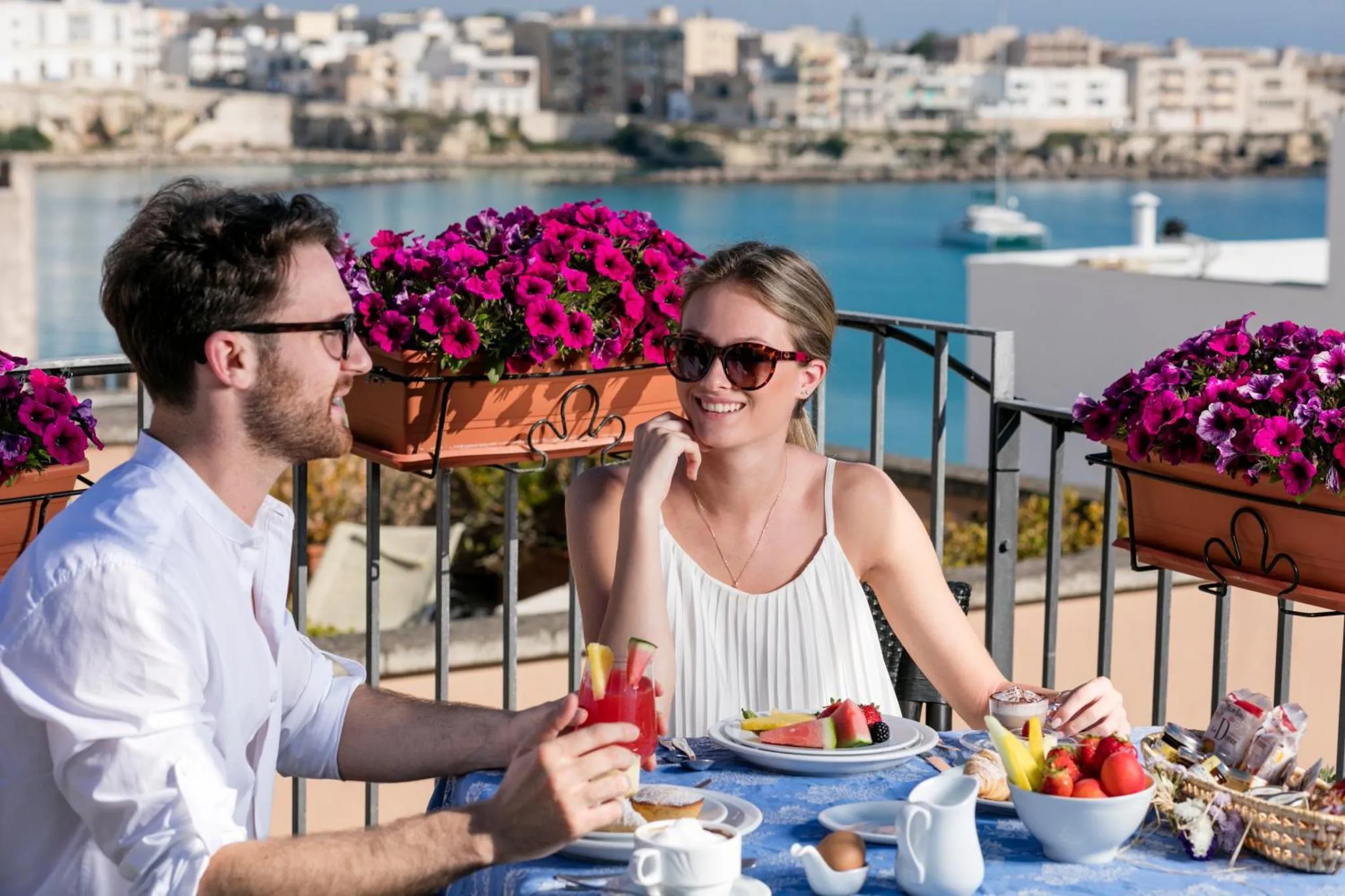 Un couple souriant partage un petit-déjeuner sur une terrasse ensoleillée avec vue sur la mer.