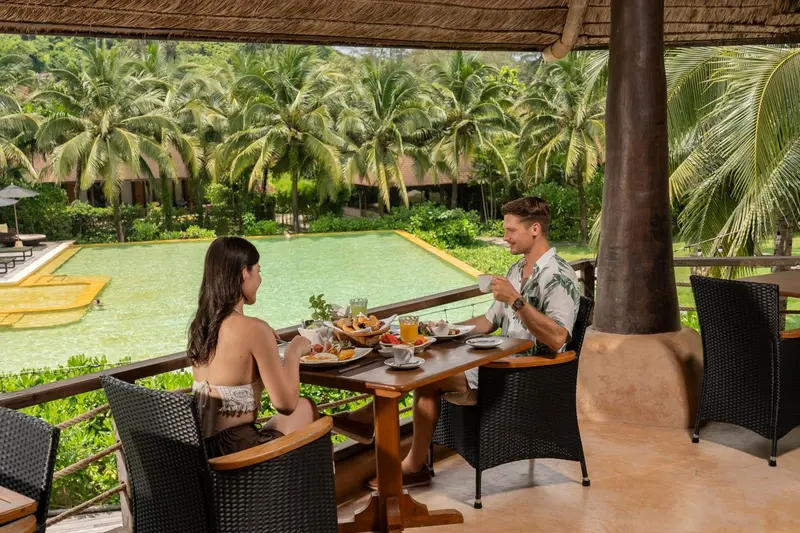 Un couple prend son petit-déjeuner à une table en osier sur une terrasse ombragée.