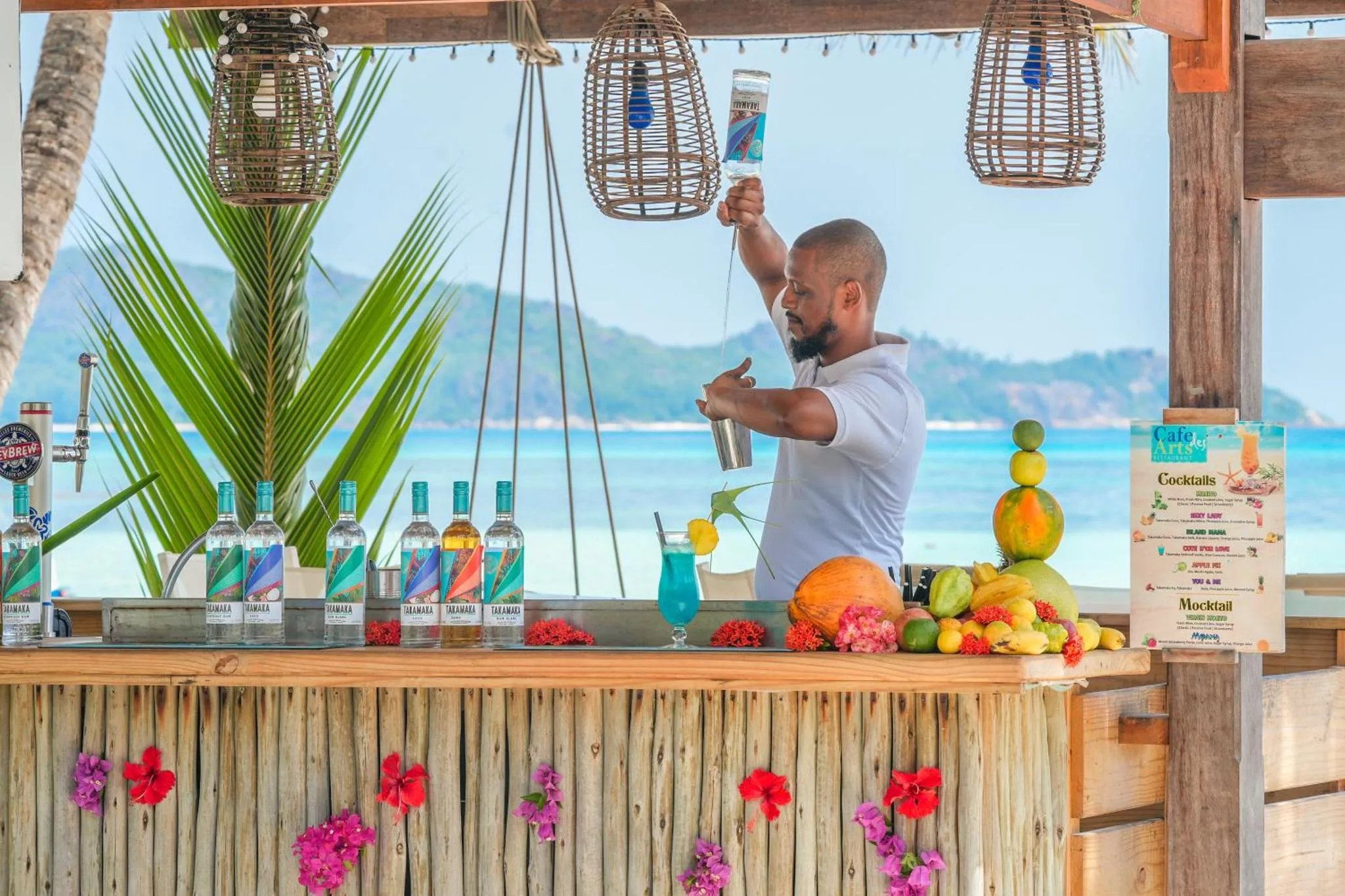 Un barman prépare un cocktail devant un comptoir décoré de fleurs tropicales, avec vue sur la mer.