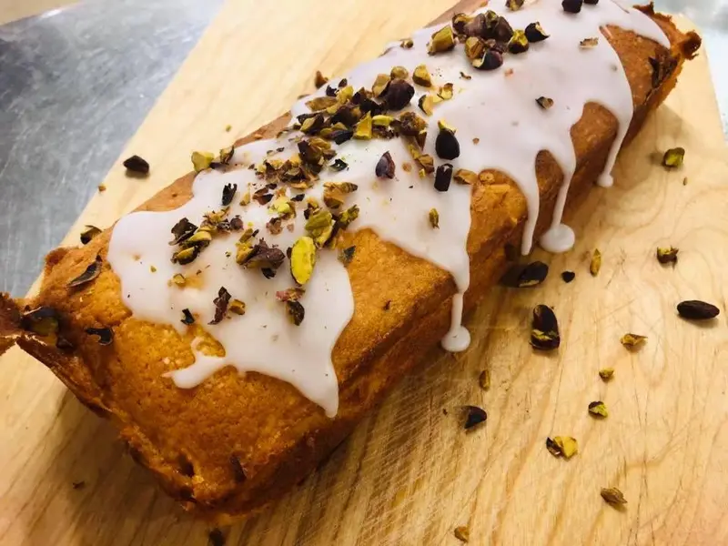 Un gâteau recouvert de glaçage blanc et de pistaches concassées est présenté sur une planche à découper en bois.