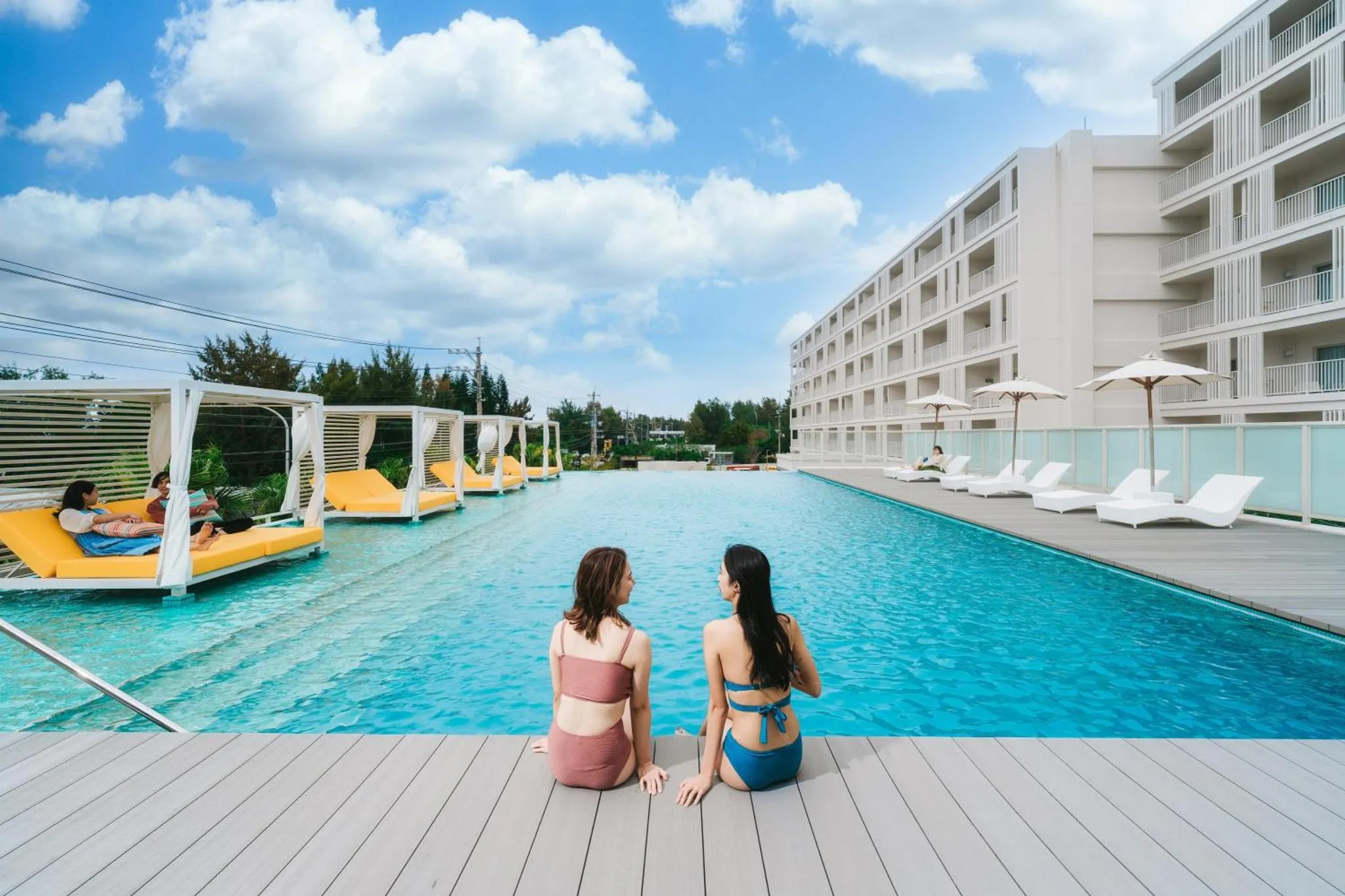 Deux femmes sont assises au bord d'une piscine rectangulaire avec des transats.