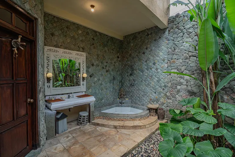Salle de bain ouverte avec baignoire et lavabo sur fond de mur en pierre.