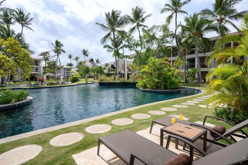 Transats et table basse avec boissons au bord d'une piscine rectangulaire entourée de végétation tropicale.