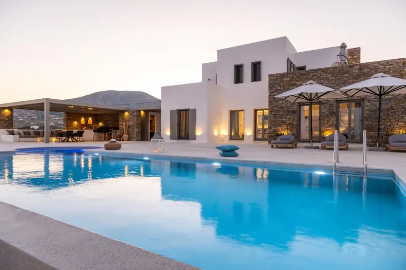 Une piscine turquoise reflète les lumières d'une villa blanche au coucher du soleil.