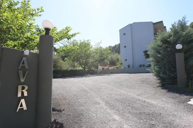 Allée gravillonnée menant à un bâtiment blanc à plusieurs étages, bordée de lampadaires et de végétation.